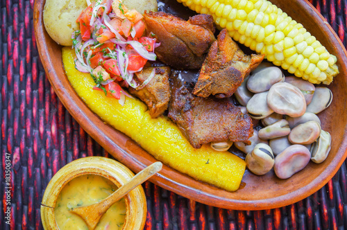 Close up of delicious hornado, ecuadorian traditional typical andean food served with corn, potato, salad, plantain and broad beans in a clay plate over a table