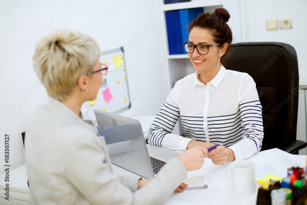 Obraz premium Smiling attractive middle aged successful financial assistant woman with eyeglasses sitting in front of the woman customer and taking a bank card in the office.