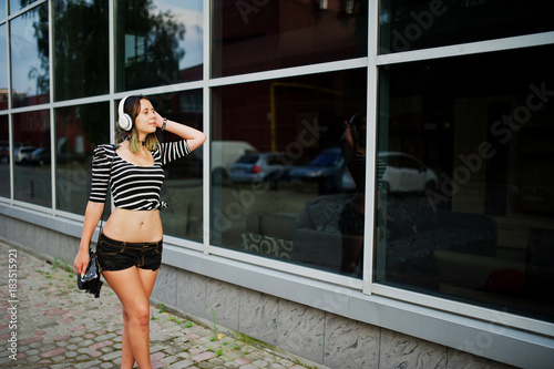 Girl wear on shorts with big headphones against large windows bulding.