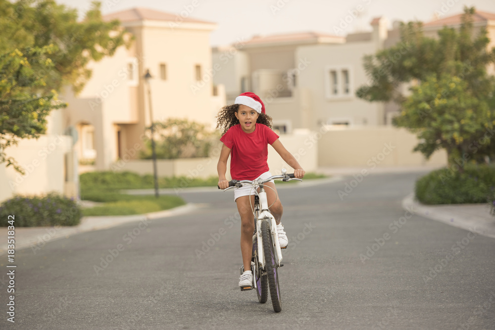 A biracial child wearing Santa hats while riding her bike on a street in a neighbourhood with homes in the background