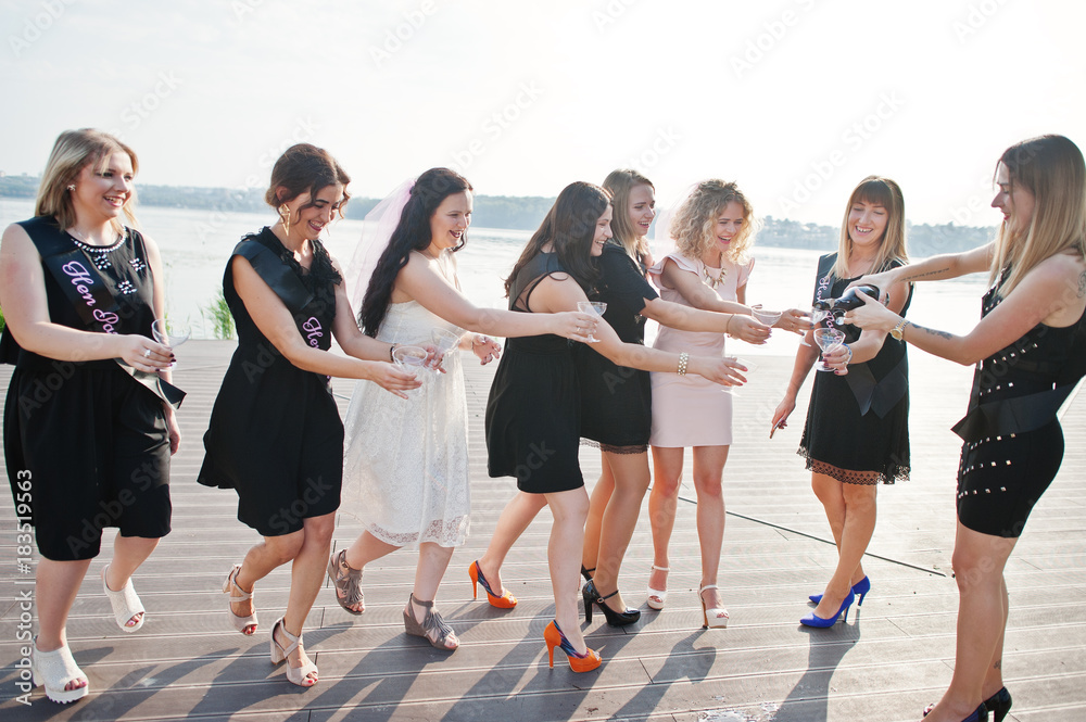 Obraz premium Group of 8 girls wear on black and 2 brides at hen party against sunny beach drinking champagne.