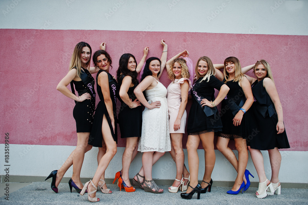 Fototapeta premium Group of 8 girls wear on black and 2 brides at hen party posed against pink wall.