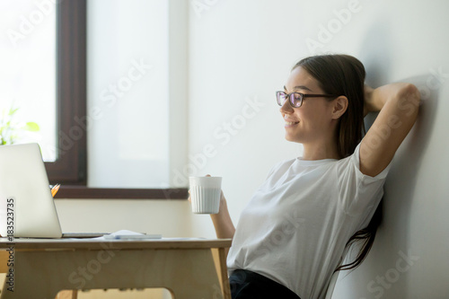 Satisfied young businesswoman holding hand behind head and drinking coffee. Beautiful female manager having break, resting and smiling photo