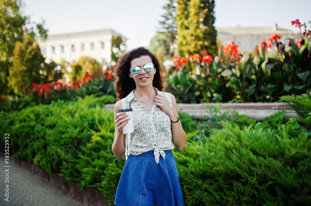 Curly stylish girl wear on blue jeans skirt, blouse and glasses posed at street of city with cup of coffee. Curly stylish girl wear on blue jeans skirt, blouse and glasses posed at street of city with cup of coffee.