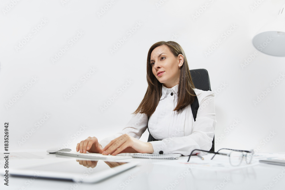 Beautiful smiling brown-hair business woman in suit and glasses sitting at the desk, working at computer with modern monitor with documents in light office, hands on keyboard, on white background Beautiful smiling brown-hair business woman in suit and glasses sitting at the desk, working at computer with modern monitor with documents in light office, hands on keyboard, on white background