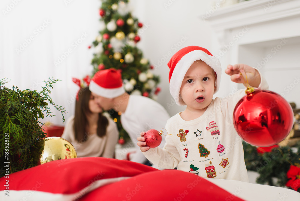 Happy young parents with cute little son in hat. Child boy with big red tree toy ball standing in decorated New Year light room at home. Christmas good mood. Family, love and holiday 2018 concept.