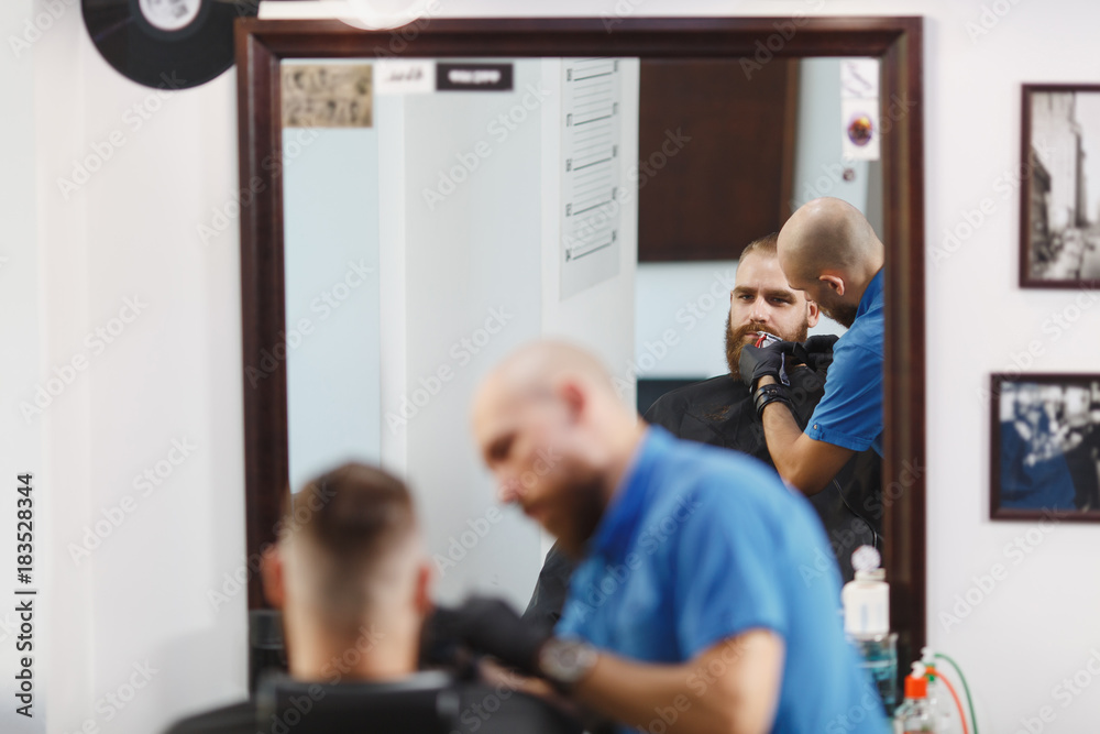Fototapeta premium Male professional hairdresser serving client, shaving thick big beard straight razor. Ginger handsome stylish young man with short hair getting trendy haircut, black cape. Light white barber shop room