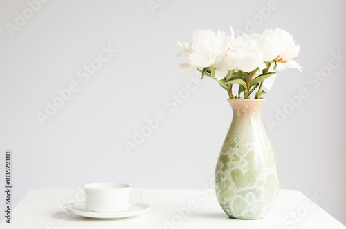 White peonies in green vase on table with cup and saucer against grey background