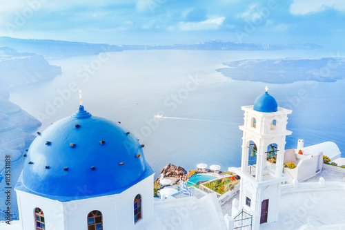 Fototapeta Grecja, wyspa Santorini w Morzu Egejskim. Zapierająca dech w piersiach sceneria z niebieskim kopulastym kościołem na pierwszym planie i epicką panoramą wyspy w tle.