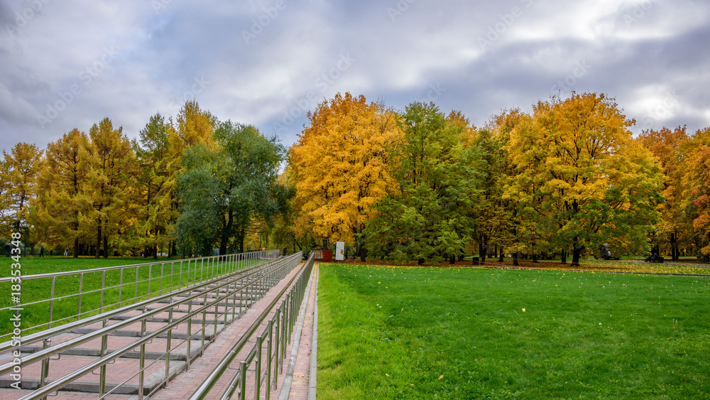 View of colorful alley with bright green and yellow trees, green lawn with dry autumn leaves on it, staircase with handrails and overcast sky in the park at autumn cold day in Moscow city, Russia