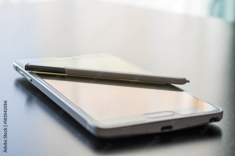 A white cellphone and pen stylus are set on a dark shiny desktop surface. Natural light is flooding into the image which creates a bright and simple yet sleek image. A white cellphone and pen stylus are set on a dark shiny desktop surface. Natural light is flooding into the image which creates a bright and simple yet sleek image.