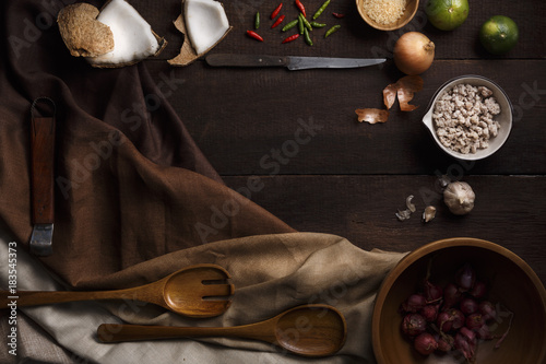 Thai food cooking ingredients with garlic, lemon, coconut, sugar, pork, chilli, wooden spoon and folk and knife on wooden board background. Top view with copy space in center