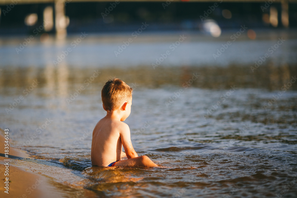 Fototapeta premium The baby boy sits on the waist in the reservoir of the river with his back and looks beyond the horizon. Concept of summer beach vacation