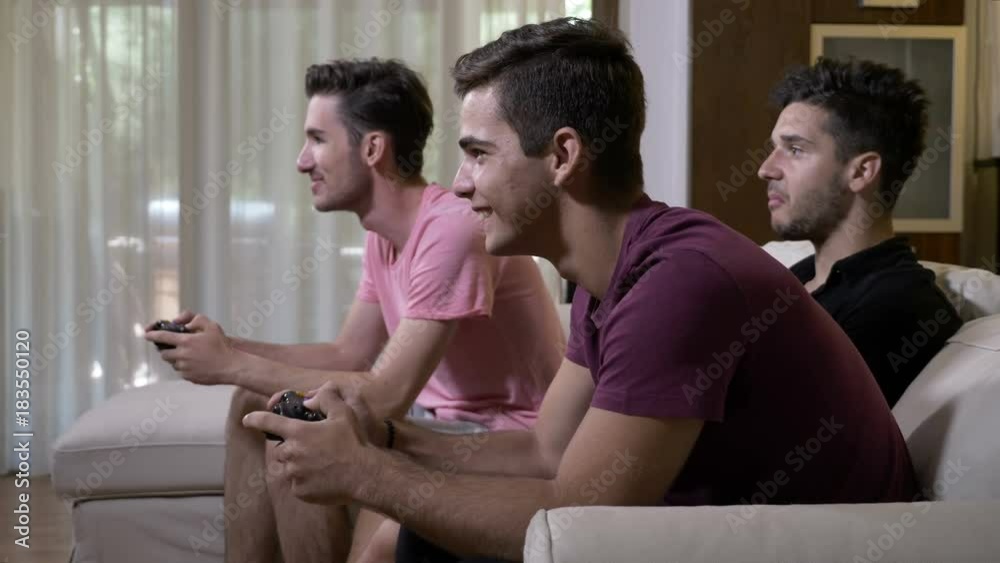 Playful teen boys sitting on couch at home playing against each other racing game on console using game joystick cheering and having fun