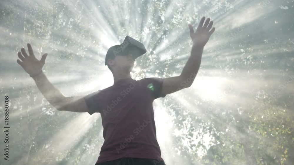 Young man with VR headset gadget immersed into virtual reality covered by light rays and waving smoke in slow motion