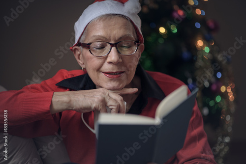 Senior old lady reading her favorite book during December holiday, house decorated with a christmas tree, she's wearing a christmas hat