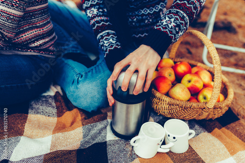 A guy opens a thermos and a girl in jeans and sweater, sitting on a blanket. Travelers pouring tea from thermos cup, outdoors. Young woman and man drinking tea at cup. Theme travel.Tonted photo. photo