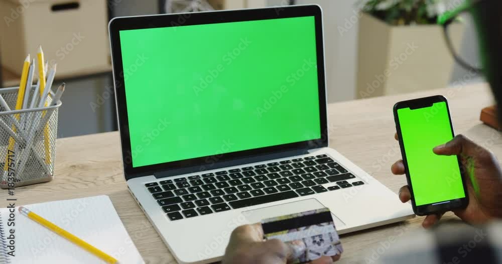 Over shoulder view on the African American man's hands scrolling and taping on a black smartphone with green screen while he holding a credit card while shopping online. Beside a laptop computer with Over shoulder view on the African American man's hands scrolling and taping on a black smartphone with green screen while he holding a credit card while shopping online. Beside a laptop computer with
