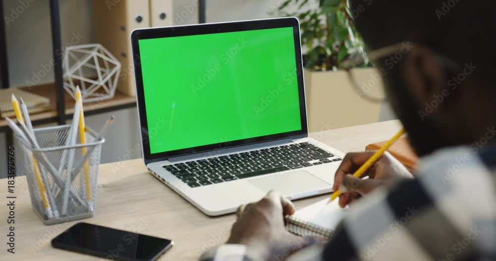 Close up of the shoulder view on the African American man writing in the notebook with a pencil and the laptop computer with a green screen and smartphone is in front him. Office room. Chroma key
