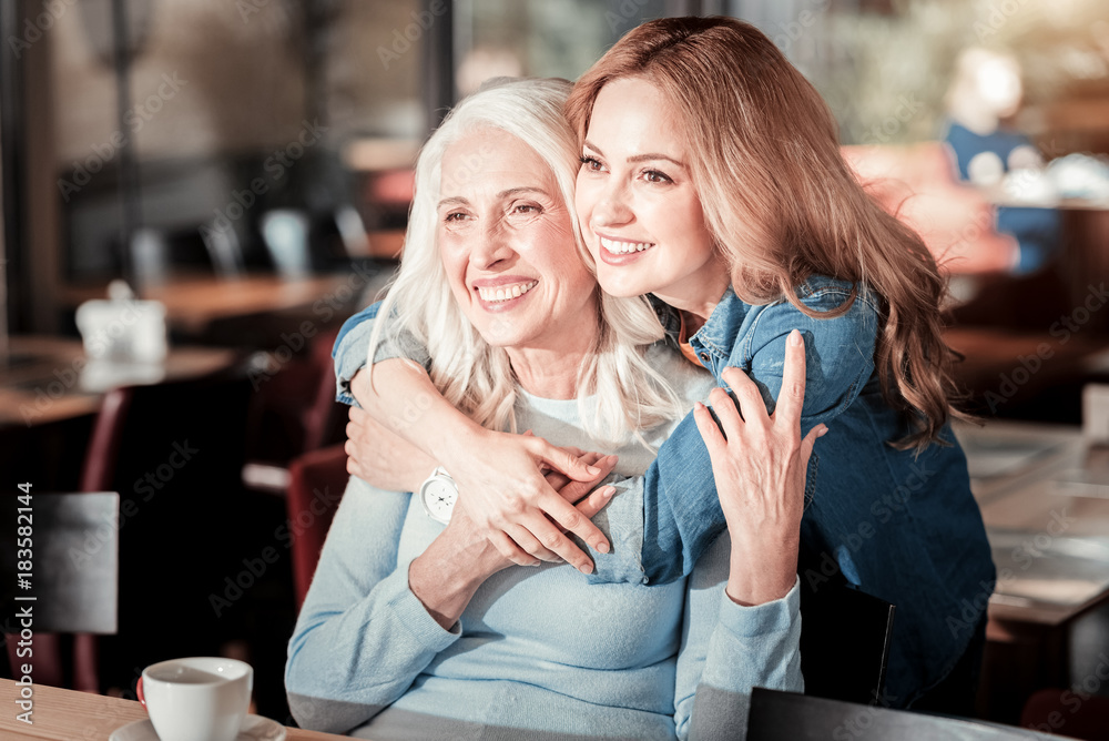 Pleasant timespending. Cheerful smart senior woman sitting and smiling while a loving attentive granddaughter standing behind her back and hugging her