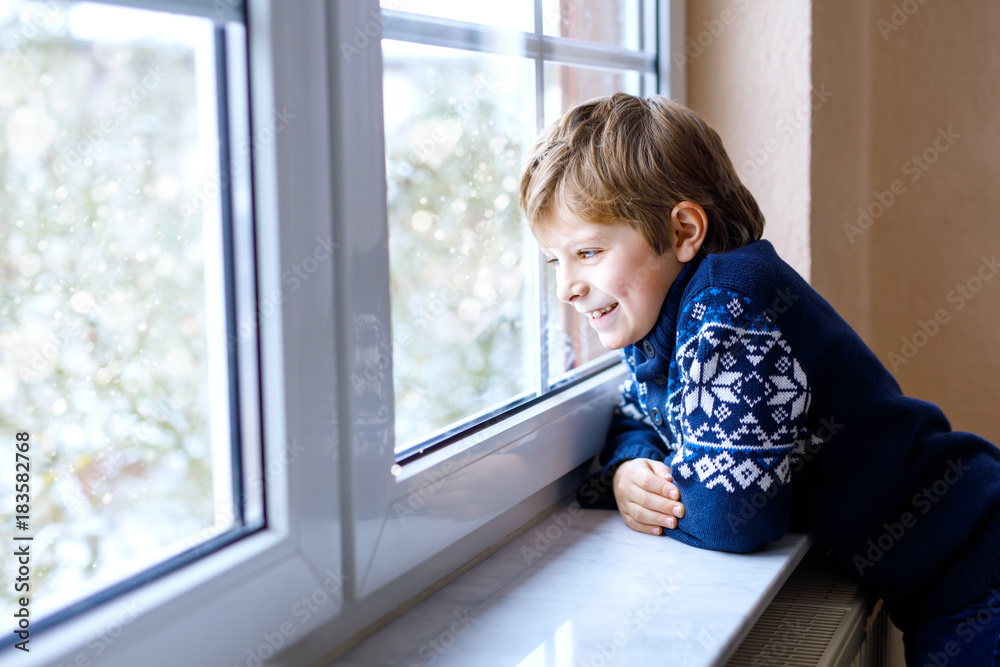 Happy adorable kid boy sitting near window and looking outside on snow on Christmas day or morning.