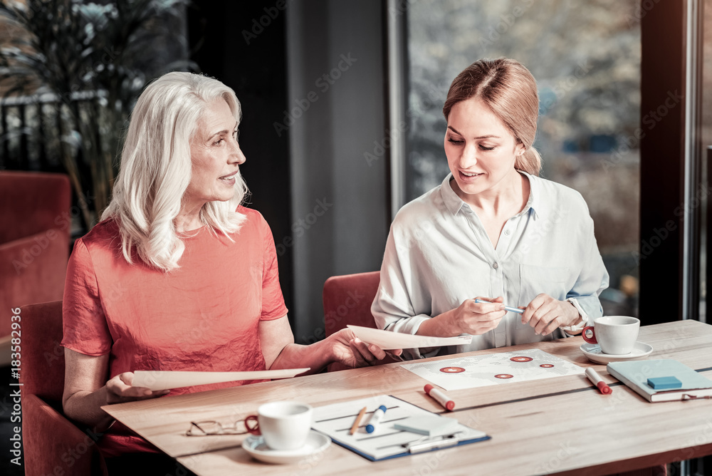 Fototapeta premium Busy women. Cheerful clever friendly designers sitting in a cafe and discussing their new exciting project with color schemes and graphics being on the table in front of them