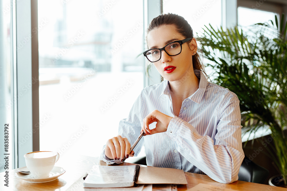 Obraz premium the business lady's portrait in office shirt and strict glasses. the woman sits at a table in cafe and makes entries in a notebook.