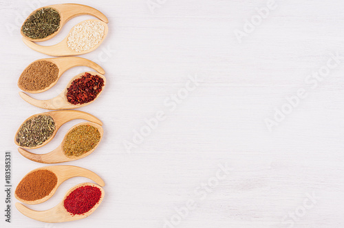 Assortment colorful powdered spices in bamboo spoons as decorative border. White wood background, top view, copy space.