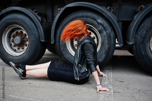 Red haired stylish girl wear in black  sitting against large truck wheels.
