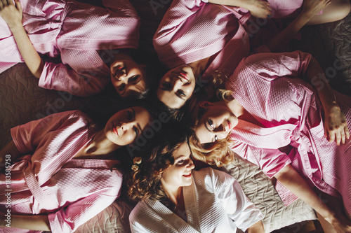Pretty bride and laughing bridesmaids in pink robes lie head to head on a bed