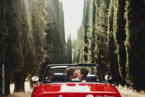 Look from behind at wedding couple kissing in a red cabrio which stands between tall trees somewhere in Tuscany, Italy