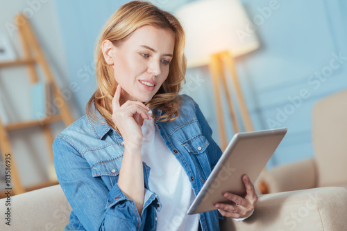 Curious. Calm concentrated young woman being attentive while sitting on a sofa in her living room and looking at the screen of a tablet with her fingers touching the face