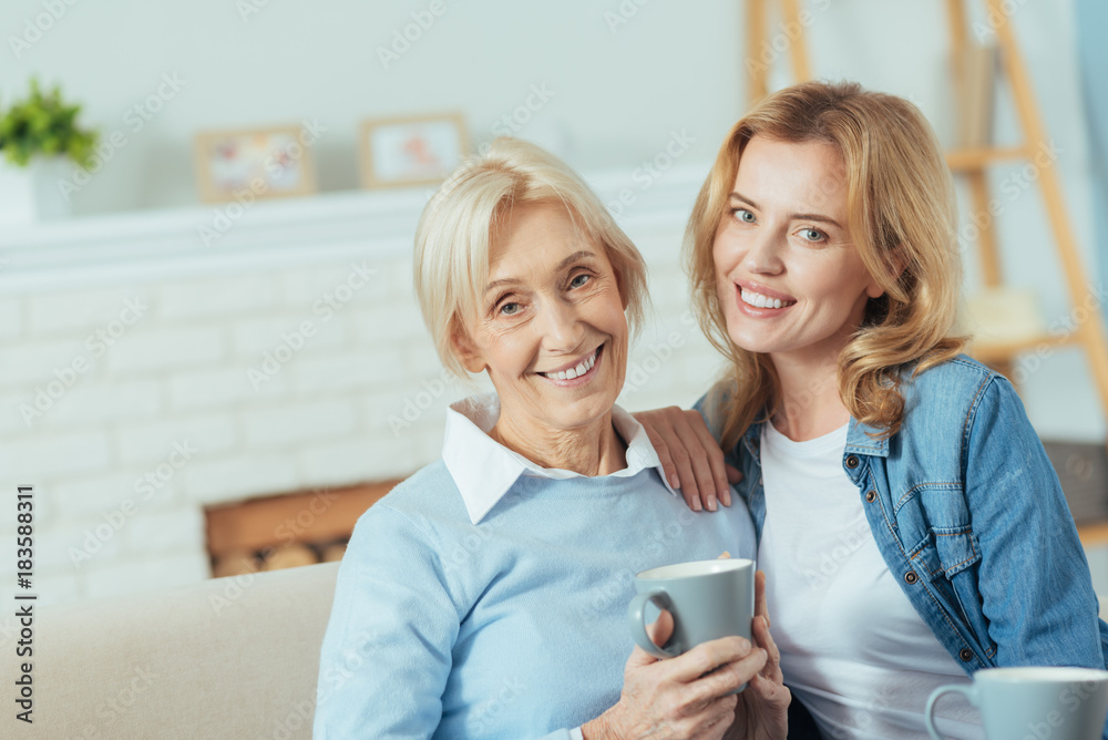 Positive relatives. Kind relaxed aged woman sitting on a soft sofa and smiling while her attentive caring granddaughter sitting by her side and putting her hand on a shoulder of a grandmother Positive relatives. Kind relaxed aged woman sitting on a soft sofa and smiling while her attentive caring granddaughter sitting by her side and putting her hand on a shoulder of a grandmother