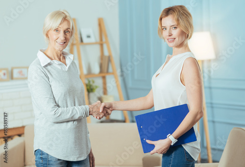 Good team. Experienced clever professional financial advisor looking confident while standing in a comfortable room and shaking hands with her pleasant cheerful aged client