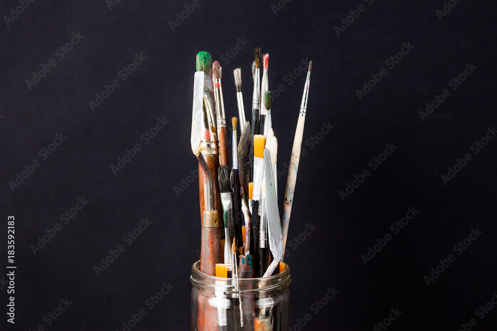 Paint brush for drawing and palette knife, dirty, in paint, in a glass, next to the white canvas. Оn a black background. With copy space. Paint brush for drawing and palette knife, dirty, in paint, in a glass, next to the white canvas. Оn a black background. With copy space.