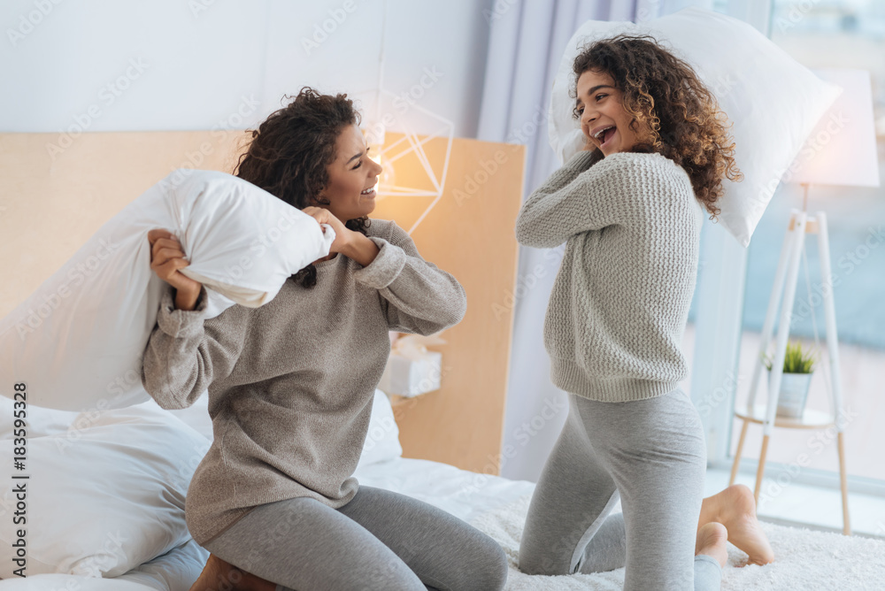 Fototapeta premium Never bored together. Positive minded young ladies beaming while playing during a leisure time and fighting with pillows on a bed.