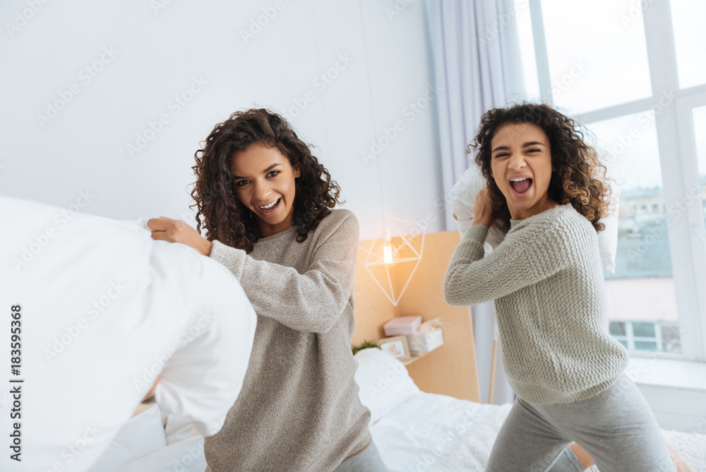 Time for a pillow fight. Joyful girls looking into the camera and grimacing while having fun and fighting with pillows together.