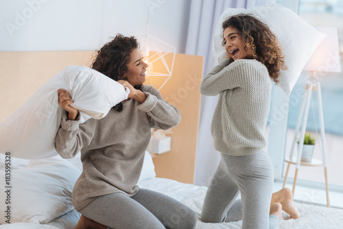 Never bored together. Positive minded young ladies beaming while playing during a leisure time and fighting with pillows on a bed.