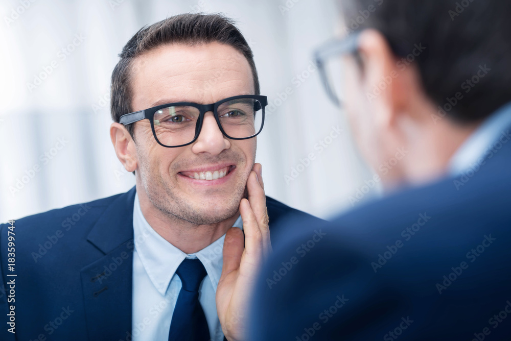 Perfect style. Optimistic budding thoughtful man touching his chick while glancing at the mirror and smiling Perfect style. Optimistic budding thoughtful man touching his chick while glancing at the mirror and smiling