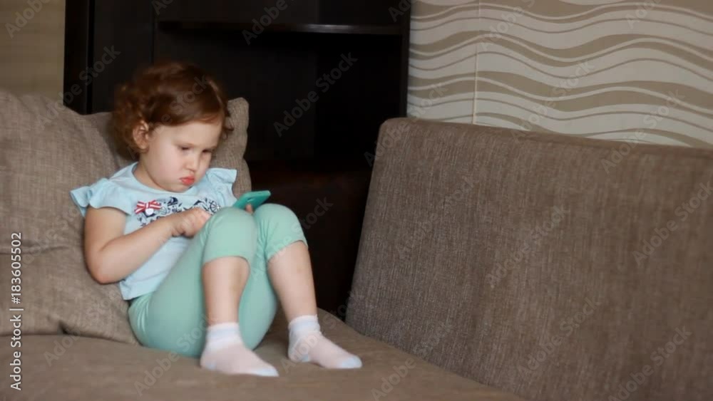Funny child looks at the phone screen and plays downloaded application on a smart phone close-up. A little cute girl lies in sofa in a living room, looking cartoon and playing the game Funny child looks at the phone screen and plays downloaded application on a smart phone close-up. A little cute girl lies in sofa in a living room, looking cartoon and playing the game
