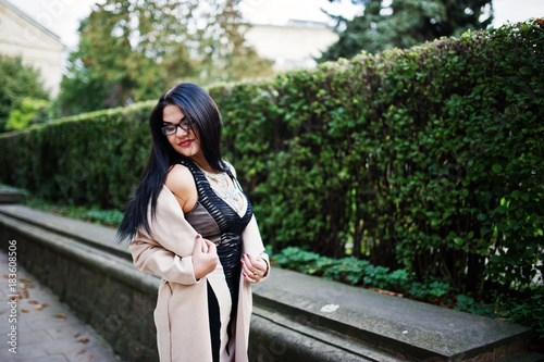 Black hair sexy woman in glasses and coat posed on street.