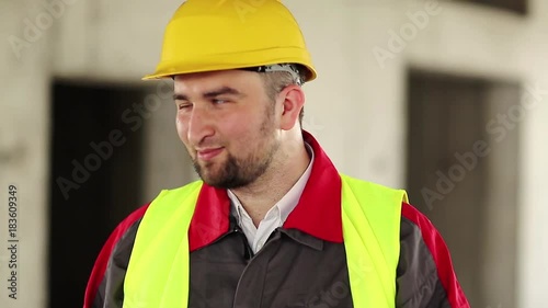 Funny builder on construction site make faces. Worker in hard hat stands on project site, looks at the camera and makes grimaces