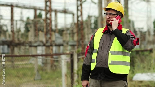 Workman at electric power plant speaks on smartphone. Power engineering specialist with cell phone and portable radio transmitter in hands on electric power station