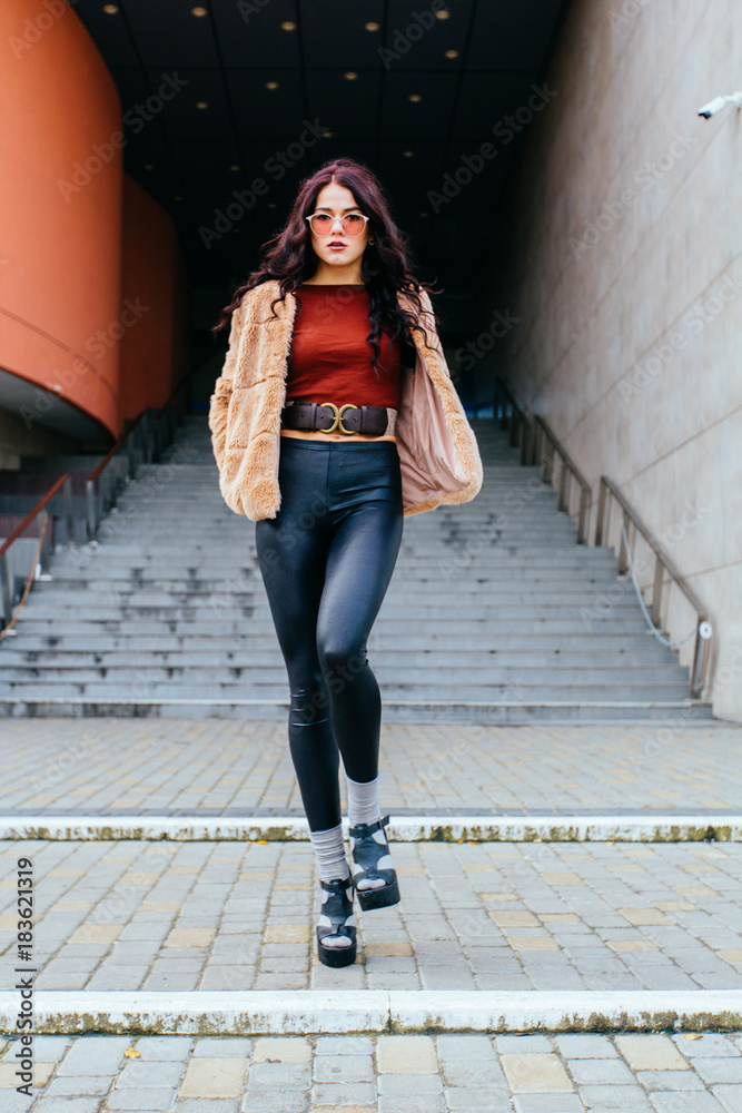 Fashion portrait of stylish sexy beautiful hippie woman in glasses model in winter warm trendy hipster clothes posing in the street over stairs background.