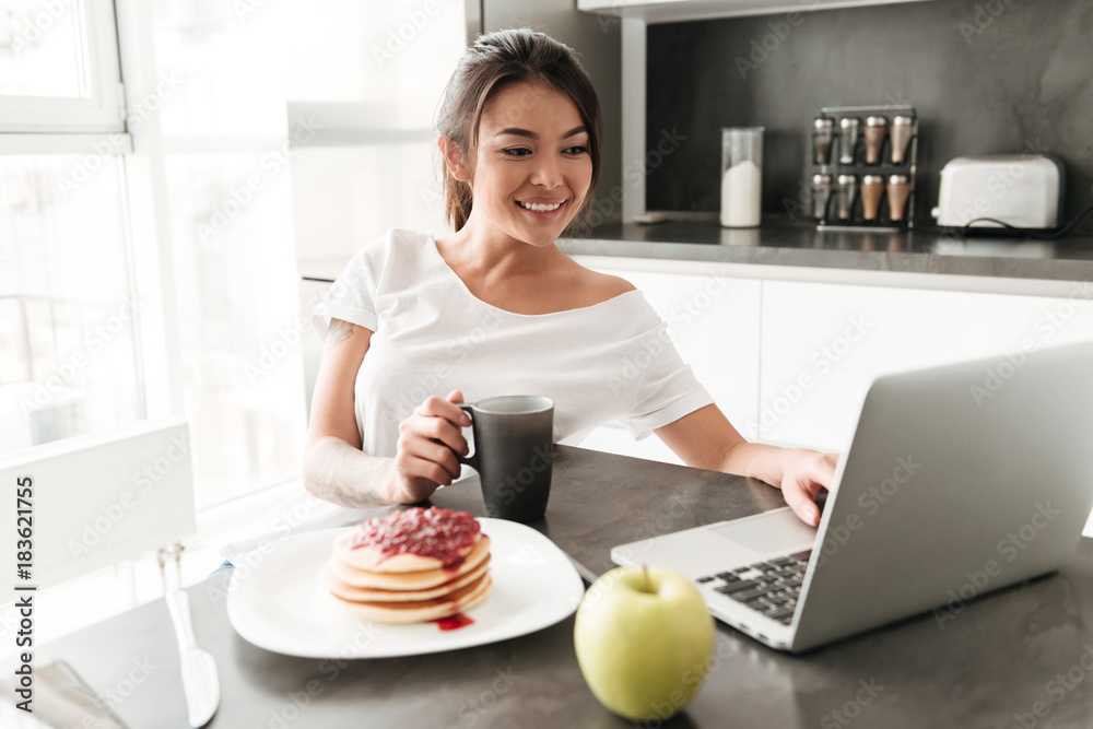 Cheerful young woman sitting at the kitchen using laptop