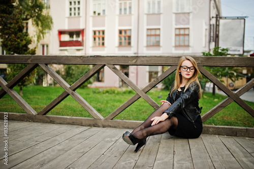 Elegant blonde girl wear on black leather jacket posing at streets of town sitting on wooden.