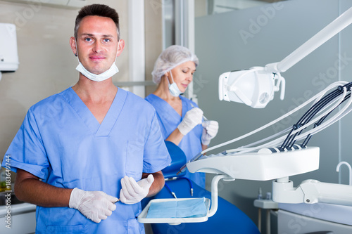 Professional smiling dentist standing in office with dental assistant background