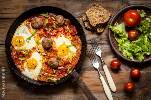 shakshuka, the Israeli dish of eggs, tomatoes and meatballs with greens on wooden background