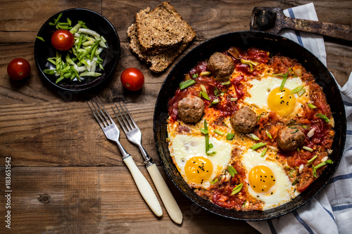shakshuka, the Israeli dish of eggs, tomatoes and meatballs with greens on wooden background