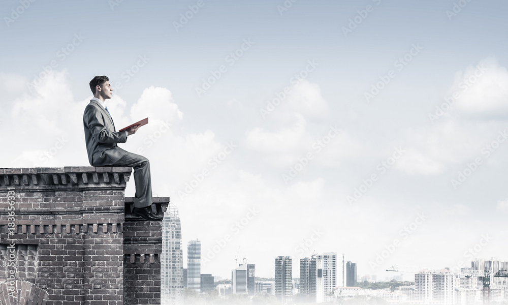 Man on roof edge reading book and cityscape at background
